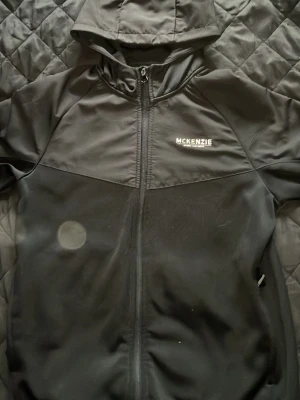 Svart hoodie från McKenzie - Snygg svart hoodie från McKenzie med dragkedja framtill och huva. Tillverkad i ett mjukt och stretchigt material som känns bekvämt. Loggan är tryckt på bröstet och modellen har långa ärmar samt ribbade muddar. Perfekt för en avslappnad streetwear-look.