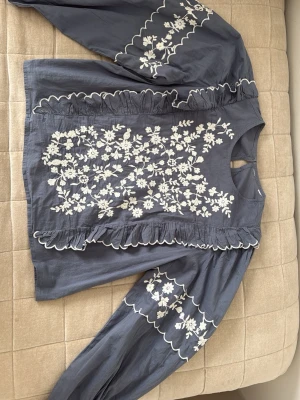 Blå blus med broderade blommor - Supersnygg blå blus med vita broderade blommor och volangdetaljer framtill. Blusen har långa, puffiga ärmar med broderier och vågiga kanter. 