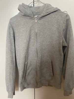 Grå hoodie från lager 157 - Ljusgrå hoodie från 157 med dragkedja framtill och två fickor. Tillverkad i mjukt sweatshirtmaterial med borstad insida för extra komfort. Klassisk huva med dragsko och ribbade muddar vid ärmslut och nederkant. Perfekt för en avslappnad och stilren look.