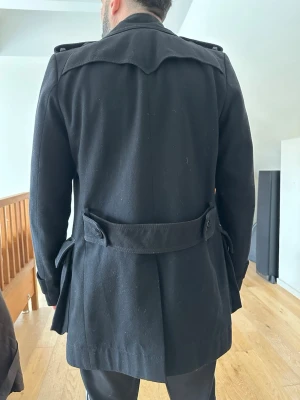 Svart trenchcoat med bälte  - Stilren svart trenchcoat med klassisk dubbelknäppning, axelklaffar och bälte i midjan. Jackan har stora fickor och en bred krage. Materialet ser ut att vara ullblandning för extra värme och struktur. Perfekt för dig som vill ha en tidlös och snygg look.