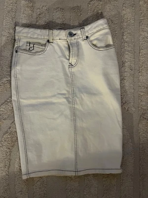 Vit vintage jeanskjol med kontrastsömmar - Säljer en vit vintage jeanskjol med markerade svarta sömmar och klassiska fem fickor. Kjolen har knapp och dragkedja framtill samt raka linjer för en clean look. Perfekt för dig som gillar enkel men snygg stil.