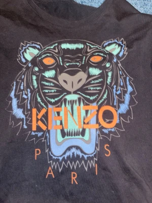 Brun långärmad topp från Kenzo - Cool brun långärmad topp från Kenzo med ikoniskt tigertryck i blått, orange och grönt på bröstet. Stora Kenzo Paris-loggan i orange framtill. Perfekt för dig som gillar statement-plagg och streetstyle.