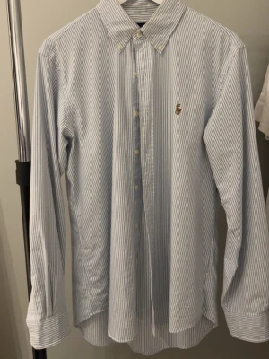 Randig skjorta från Polo Ralph Lauren - Klassisk blå- och vitrandig skjorta från Polo Ralph Lauren med broderad logga på bröstet. Skjortan har lång ärm, knappar framtill och button-down krage. Perfekt för en stilren look.