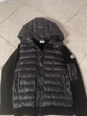 Svart dunjacka med huva från Moncler - Snygg svart dunjacka från Moncler med quiltad front och huva. Ärmarna är i stickat material och jackan har dragkedja framtill samt två fickor. Moncler-logga på ena ärmen. Perfekt för kalla vinterdagar. Pris kan diskuteras