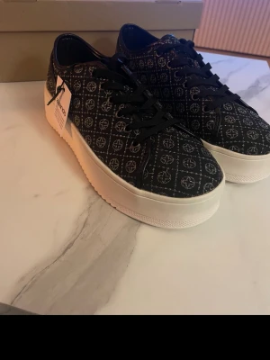 Anna Field sneakers med mönster - Snygga svarta sneakers från Anna Field med vitt mönster och klassisk logga. Skorna har svarta snören och en vit, tjock sula som ger en modern look. Materialet är textil med detaljer i syntet. Perfekta för dig som vill sticka ut med stil.