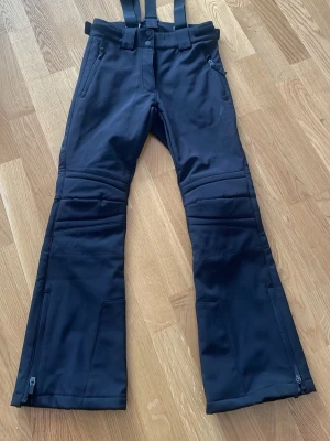 Svarta täckbyxor med avtagbara hängslen - Säljer ett par svarta täckbyxor med justerbara hängslen samt att det går att justera i midjan och dragkedjor vid bensluten. Byxorna har två framfickor med dragkedja och förstärkta knän. Perfekta för kalla dagar i backen eller snölek.