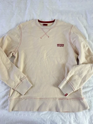 Beige sweatshirt från Levi's - Säljer en beige sweatshirt från Levi's med röda detaljer och logga på bröstet. Tröjan har rund halsringning, ribbade muddar och sömmar i kontrastfärg. Perfekt för en avslappnad och stilren look.