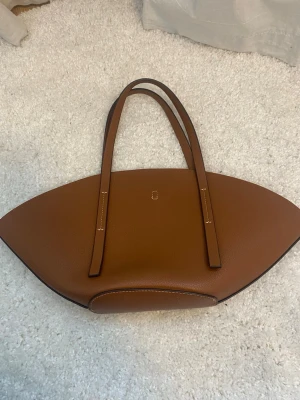 Brun handväska GinaTricot  - Säljer min supersnygga väska från Gina Tricot i nyskick. Väskan har en unik, bågformad design med två långa handtag och vita kontrastsömmar. Insidan är rymlig och har metallhake. Hör av dig vid frågor!💛