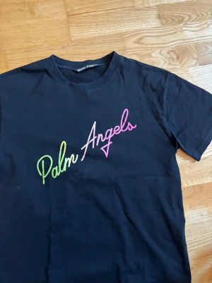 Svart Palm Angels t-shirt - Svart t-shirt från Palm Angels med färgglad logotyp i grönt och rosa på bröstet. Klassisk rund halsringning och korta ärmar. Perfekt för en trendig streetwear-look.