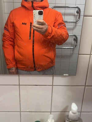 Orange pufferjacka från Helly Hansen - Säljer en riktigt fet orange pufferjacka från Helly Hansen med svart dragkedja och logga på bröstet. Jackan har huva och två sidofickor, samt coola detaljer på ärmarna. Perfekt för kalla dagar när du vill sticka ut.