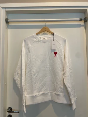 Vit sweatshirt från Ami Paris - Vit sweatshirt från Ami Paris med röd broderad hjärtdetalj och bokstaven A på bröstet. Tröjan har rund halsringning och långa ärmar. Perfekt för en avslappnad och stilren look.
