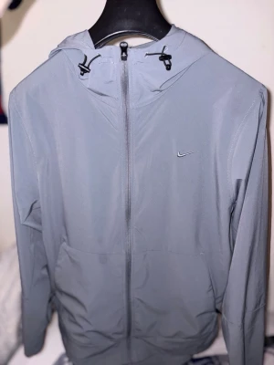 Grå Nike hoodie med dragkedja - Snygg grå hoodie från Nike med dragkedja framtill och justerbar huva med snören. Tröjan har fickor på sidorna och en liten Nike-logga på bröstet. Perfekt för träning eller chill. Tillverkad i mjukt och lätt material.