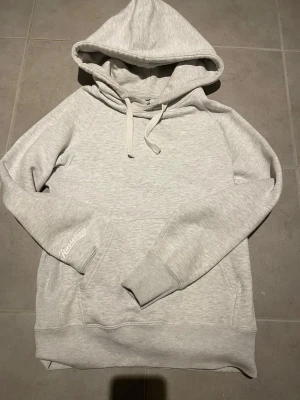 Ljusgrå hoodie från Russetress - Snygg ljusgrå hoodie från Russetress med klassisk huva och dragsko. Hoodien har ribbade muddar vid ärmslut och nederkant samt en stor magficka. Tillverkad i mjukt bomullsmaterial och har Russetress-logga broderad på ena ärmen.