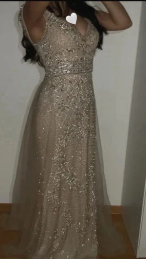 Glittrig långklänning i champagne - Säljer en magisk långklänning i champagnefärg med glittriga paljettdetaljer över hela klänningen. Klänningen har en elegant, figurnära överdel och ett svepande, transparent tyg i kjolen. Perfekt för bal eller festliga tillfällen.