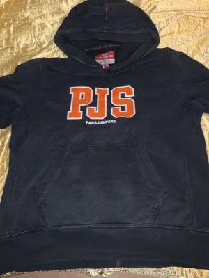 Svart hoodie från Parajumpers - Svart hoodie från Parajumpers med stor orange PJS-logga på bröstet och broderad patch på ärmen. Klassisk känguruficka framtill och huva med röda sömmar. Tillverkad i mjuk fleece, perfekt för en avslappnad streetwear-look. Tröjan har tappat ganska mycket färg men den är fortfarande fin och värt priset enligt mig!