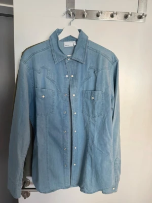 Ljusblå jeansjacka från ASOS DESIGN - Säljer en ljusblå jeansjacka från ASOS DESIGN med western-inspirerade sömmar, två bröstfickor med knappar och tryckknappar framtill. Klassisk krage och raka ärmar. Perfekt för dig som gillar en avslappnad och trendig look.