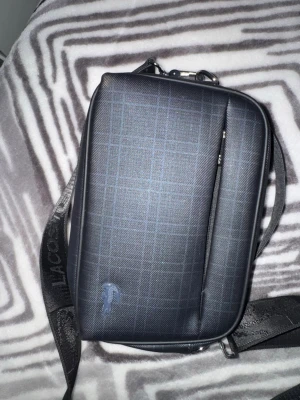 Rutigt axelväska från Lacoste - Snygg axelväska från Lacoste i svart med diskret rutmönster och blå logga. Väskan har justerbar axelrem med Lacoste-tryck, dragkedjefack framtill och metallplatta med logotyp. Perfekt för att bära det viktigaste med stil. Pris kan diskuteras 