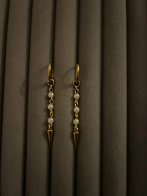 Guldiga örhängen med pärlor och spets - Snygga örhängen i guldfärgad metall med små vita pärlor på rad och en spetsig konformad detalj längst ner. Hängande modell som ger en cool och elegant vibe. Perfekta för dig som gillar unika accessoarer med lite edge.