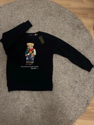 Svart tröja Polo Bear Ralph Lauren - Cool svart tröja från Ralph Lauren med Polo Bear-motiv på framsidan. Tröjan har rund hals och långa ärmar. Björnen på trycket har färgglada kläder och texten 'POLO BEAR BY RALPH LAUREN' under. Perfekt för dig som gillar ikoniska prints.