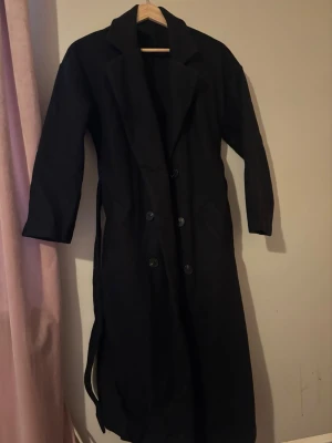 Svart lång trenchcoat med knytskärp - Stilren svart trenchcoat med dubbelknäppning och klassisk krage. Lång modell med knytskärp i midjan och två fickor framtill. 