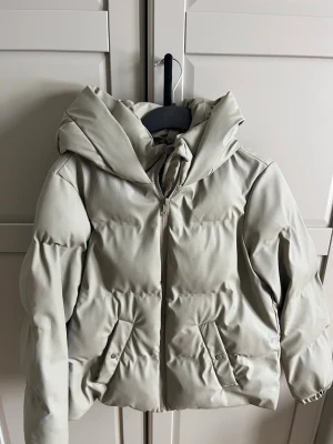 Beige pufferjacka med huva - Snygg beige pufferjacka med stor huva och dragkedja framtill. Jackan har två framfickor med knappstängning och en boxig, trendig passform. Perfekt för kalla dagar och ger en clean look. Materialet är slätt och quiltat för extra värme.