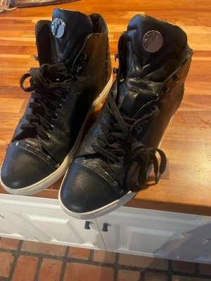 Svarta high top sneakers i läder - Snygga svarta high top sneakers från Versace i äkta läder med vita sulor och svarta skosnören. Skorna har en metalllogga på plösen och stilrena detaljer längs sidorna. Perfekta för dig som vill ha en lyxig och trendig look.