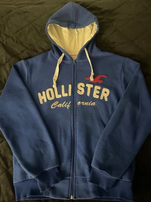 Blå zip-hoodie från Hollister - Blå hoodie med dragkedja från Hollister. Stor broderad logga och text i gult och rött framtill, samt vita snören och ljus insida på huvan. Mjuk bomullskänsla och ribbade muddar vid ärmslut och nederkant. Perfekt för en avslappnad stil. Kan gå ner i pris vid snabb affär