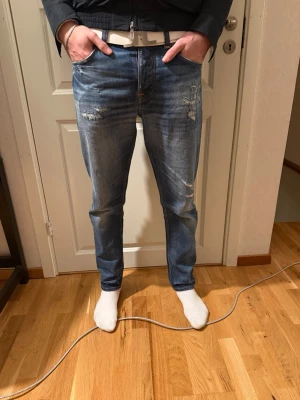 Blå slitna jeans från nudie - Säljer ett par blå jeans från nudie med snygga slitningar och tvättade detaljer. Jeansen har rak passform, klassiska fem fickor och guldfärgade dragkedjor på fickorna. Jeansen är selvedge.  Perfekta för en avslappnad och trendig look.