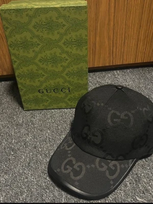 Svart keps från Gucci med GG-mönster - Snygg svart keps från Gucci med det klassiska GG-monogrammet över hela kepsen. Kepsen har böjd skärm och är tillverkad i ett tygmaterial med exklusiv känsla. Perfekt accessoar för att lyfta din streetwear-look.