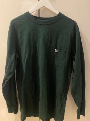 Mörkgrön långärmad t-shirt från Lee - Mörkgrön långärmad t-shirt från Lee med klassisk passform. Tröjan har en bröstficka med diskret Lee-logga och är gjord i mjuk bomull. Perfekt för en avslappnad och stilren look.
