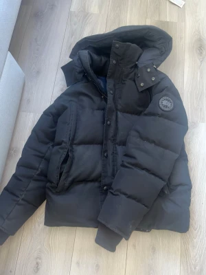 Svart dunjacka från Canada Goose - Säljer en svart dunjacka från Canada Goose med huva och patch-logga på ärmen. Jackan har stora fickor framtill, dragkedja och knappar samt en klassisk puffig design. Perfekt för kalla vinterdagar och riktigt varm.