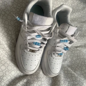 Nike Nocta Air Force 1  - Klassiska vita Nike Air Force 1 sneakers med  tå, blå detaljer på snörningen och Nike Air-logga på plös och häl. Skorna har en låg siluett och är tillverkade i slätt läder med vit sula. Perfekta för en clean och stilren look.