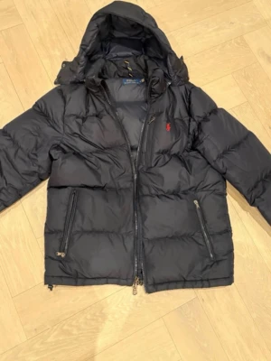 Polo ralphlauren jacka  - Svart dunjacka från Polo Ralph Lauren med huva, två dragkedjeförsedda fickor och klassisk röd logga på bröstet. Jackan är vadderad och har en sportig look med breda kanaler. Perfekt för kalla vinterdagar. Den är väldigt varm och snygg vid frågor hör av er 