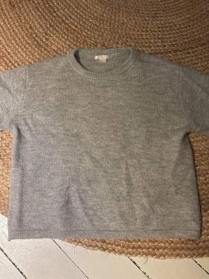Grå stickad tröja  - En stilren grå stickad tröja från COS med rund halsringning och rak passform. Tröjan har korta ärmar och är tillverkad i mjukt material som ger en clean och minimalistisk look. Perfekt för lager på lager och enkel att matcha.