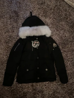 Svart dunjacka med pälskant från Moose Knuckles - Säljer en svart dunjacka från Moose Knuckles med stor vit pälskant på huvan och tydliga loggor på bröstet och ärmen. Jackan har dragkedjor på ärmarna, tryckknappar framtill och flera fickor. Perfekt för kalla vinterdagar. Förpackning finns med!