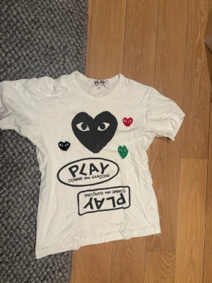 Vit Comme des Garçons PLAY t-shirt - Vit t-shirt från Comme des Garçons PLAY med ikoniska hjärtmotiv i svart, rött, grönt och svart tryck framtill. 