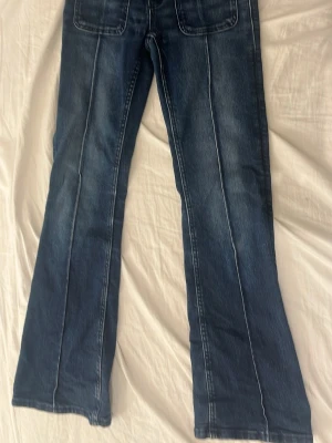 Blå bootcut jeans med fickor - Säljer ett par blå bootcut jeans med markerade sömmar och stora fickor fram och bak. Jeansen har en klassisk tvättad look och är tillverkade i denim. Perfekta för dig som gillar retrovibbar och vill ha ett par jeans med lite vidare ben.