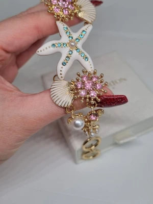 Armband med sjöstjärna och pärlor - Unikt armband med havsinspirerade detaljer som vit sjöstjärna, snäckor och blommor dekorerade med rosa och blå strass. Guldfärgad metall med pärlliknande dekorationer och justerbar kedja. Perfekt för dig som gillar statement-smycken och strandvibbar. Lådan ingår ej.