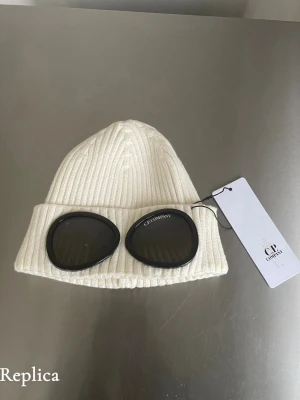Vit C.P. Company Mössa - Vit ribbstickad mössa från C.P. Company med ikoniska svarta glasögonlinser framtill. Mössan är tillverkad i mjukt material och har en uppvikt kant för extra stilpoäng. Perfekt accessoar för att sticka ut i mängden.
