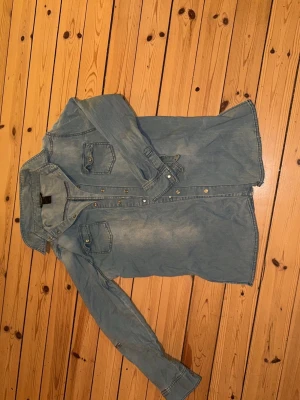 Ljusblå jeansskjorta med knappar - Snygg ljusblå jeansskjorta med klassisk krage och två bröstfickor med lock. Skjortan har långa ärmar och knäppning framtill med metallknappar. Perfekt för en avslappnad och trendig look.