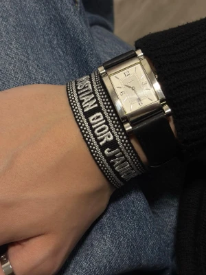 Svart broderat armband Dior - Snyggt svart och vitt broderat armband från Christian Dior med texten 'Christian Dior J'Adior'. Armbandet har justerbara band med tofsar i ändarna och är gjort i textilmaterial. Perfekt accessoar för att lyfta din outfit.
