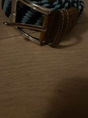 Randigt flätat bälte med skinn-detaljer - Snyggt flätat bälte i blått och svart med bruna skinn-detaljer och silverfärgat spänne. Perfekt accessoar för att lyfta din outfit med en cool och modern touch. Bältet har en klassisk design med kontrastsömmar på läderdelarna.