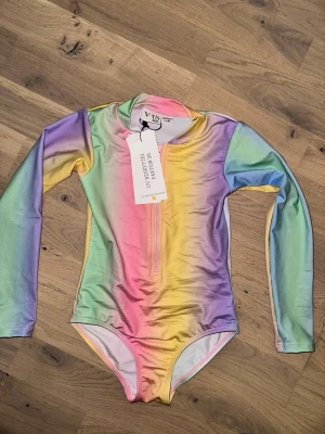 Regnbågsfärgad långärmad baddräkt VRS - Snygg långärmad baddräkt från VRS i regnbågsfärgat mönster med dragkedja framtill. Materialet är stretchigt och snabbtorkande, perfekt för strand eller pool. Färgerna går i pastelltoner av rosa, blått, gult, grönt och lila. Rund halsringning och figurnära passform.