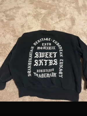Svart sweatshirt från SWEET SKTBS - Cool svart sweatshirt från SWEET SKTBS med stor vit text och grafik på ryggen. Tröjan har rund halsringning, ribbade muddar och en avslappnad passform. Perfekt för dig som gillar streetwear och vill sticka ut med snygg print.