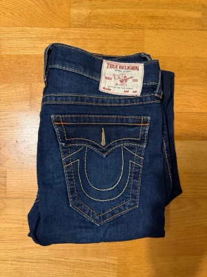 Mörkblå True Religion jeans - Säljer ett par mörkblå jeans från True Religion med klassisk kontrastsöm och stor fickdetalj bak. Jeansen har raka ben och fem fickor. Materialet är denim i bomull och passformen är avslappnad. Perfekta för dig som gillar streetwear och snygga detaljer.