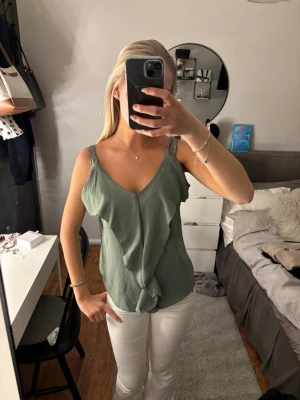 Grön volanglinne med spetsband - Snyggt grönt linne med volangdetaljer framtill och tunna axelband i spets. Djup v-ringning och avslappnad passform, perfekt för en trendig och fräsch look. Materialet känns lätt och luftigt, vilket gör toppen superbekväm.