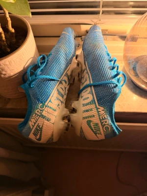  Nike Mercurial vapor 13 elite  - Säljer ett par blå Nike Mercurial fotbollsskor med vita och turkosa detaljer. Skorna har snörning, låg profil och textilöverdel för bra bollkontroll. Sulan är vit med svarta dobbar, perfekt för spel på gräs. Snygg design med Mercurial- och Nike-logga på sidan.