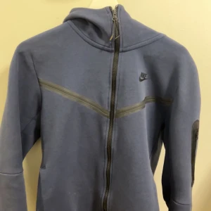 Nike tech kofta mörk blå - Snygg blå huvtröja från Nike med dragkedja och långärmade ärmar. Perfekt för en avslappnad stil eller träning. Den har en diskret Nike-logga på bröstet. Lite skada i fickan och den svarta grejen som man ser 