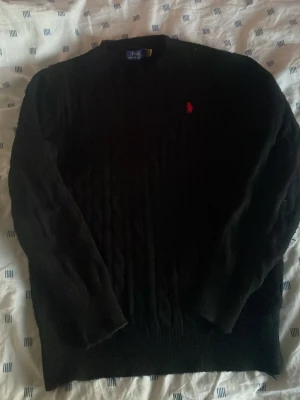Svart kabelstickad tröja Polo Ralph Lauren - Kabelstickad svart tröja från Polo Ralph Lauren med klassisk röd logga broderad på bröstet. Tröjan har rund halsringning och långa ärmar. Perfekt för dig som gillar stilrena och tidlösa plagg. Äkta och använt.