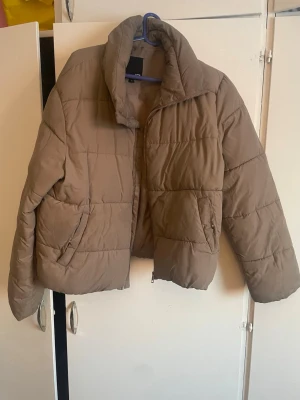 Brun pufferjacka från 157 - Säljer en brun pufferjacka från 157 med hög krage och dragkedja framtill. Jackan har en boxig passform, två sidofickor och quiltad design. Perfekt för kalla dagar och riktigt skön att bära. Materialet är syntet och jackan är fodrad för extra värme.